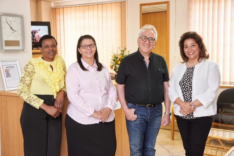 Drs. Bassem Fawzy & Tereza Fawzy, receptionist Juleen, and a patient standing next to each other, smiling, by the reception desk of the practice.